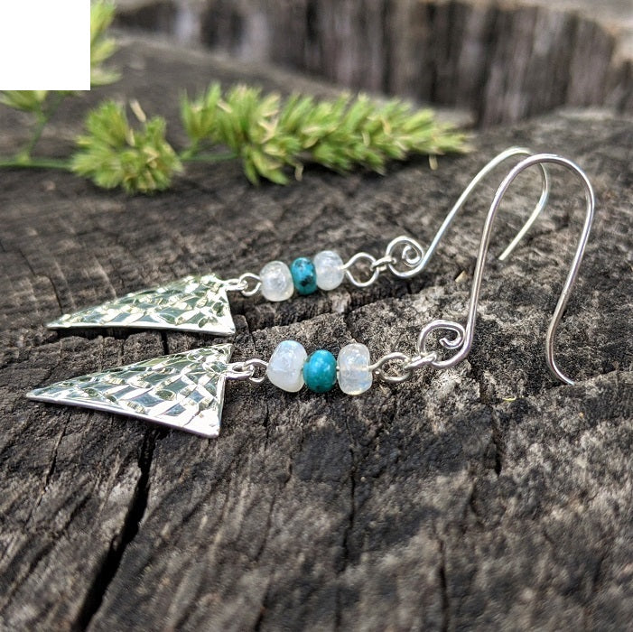 Sterling Silver Turquoise and Moonstone Arrowhead Earrings from Danare Designs lay on a rustic log