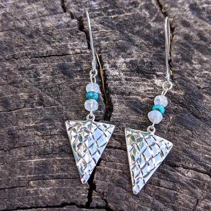 Sterling Silver Turquoise and Moonstone Arrowhead Earrings from Danare Designs lay on a rustic log
